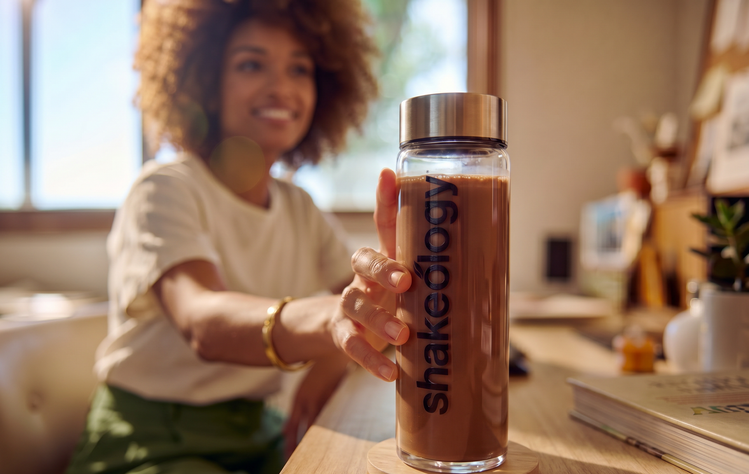 A woman in a white t-shirt and green pants smiles while holding a clear Shakeology insulated bottle filled with an iced coffee on a wooden desk in a warm, sunlit room.