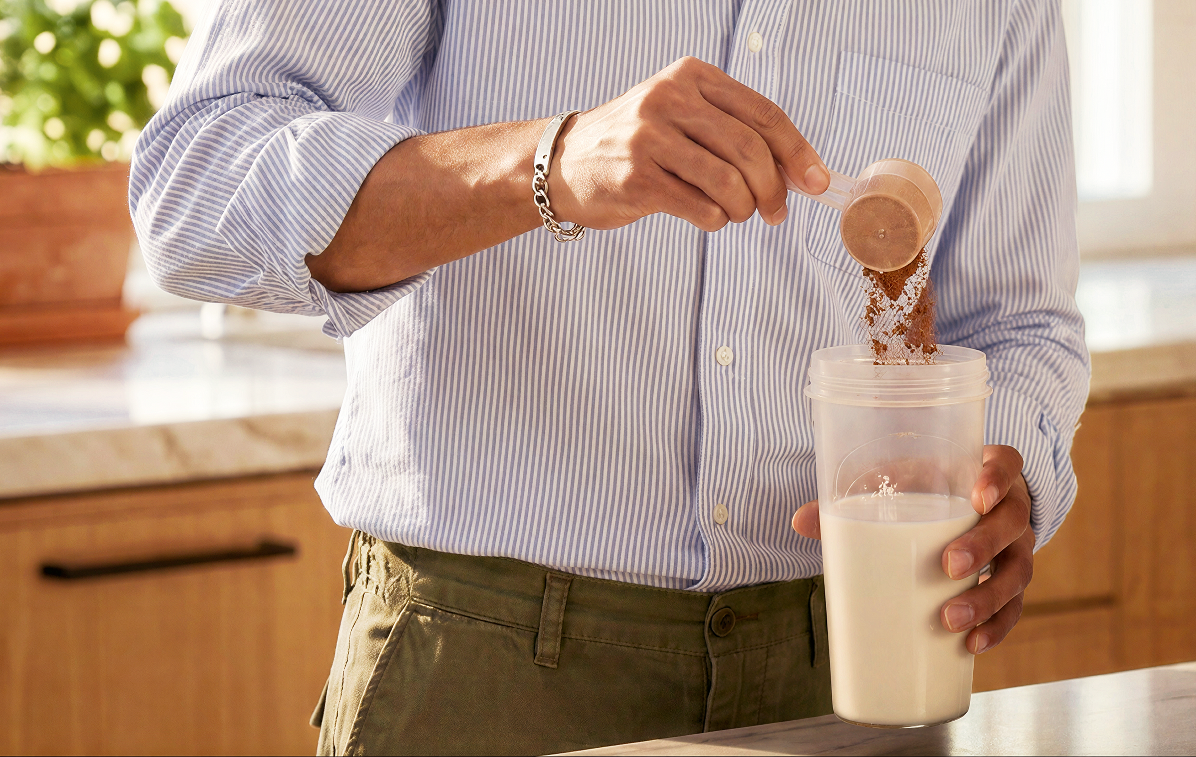 A man in a blue and white striped button-down shirt and olive green pants pours a brown powder from a wooden measuring scoop into a clear plastic shaker bottle containing milk, set in a bright kitchen environment.