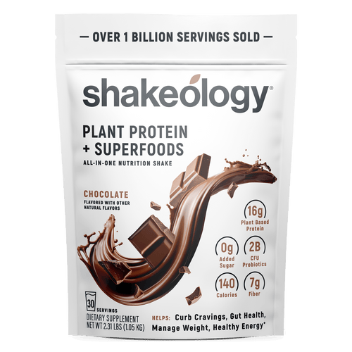 A stand-up pouch of Shakeology Plant Protein + Superfoods in chocolate flavor, shown on a plain white background. The packaging is white with black text and a large chocolate splash graphic on the front, plus callout icons for protein, added sugar, calories, and fiber.